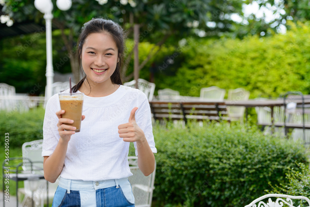 Happy young beautiful Asian woman with coffee giving thumbs up at the coffee shop outdoors