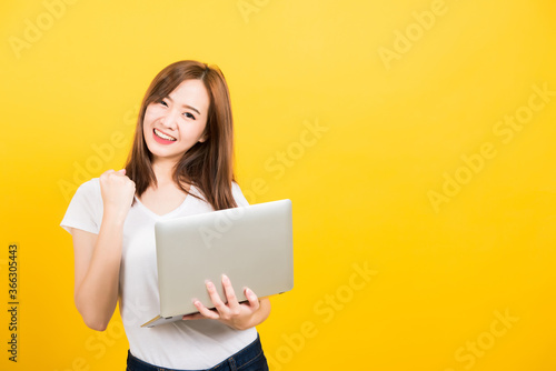 Asian happy portrait beautiful cute young woman teen smile standing wear t-shirt hold laptop computer and excited celebrating success looking to camera, studio shot yellow background with copy space