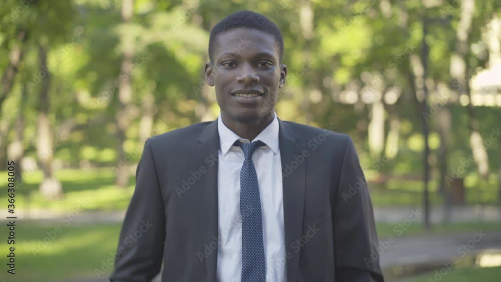 Middle shot portrait of confident businessman in suit adjusting necktie and smiling at camera. Young African American man posing in sunlight in summer park outdoors. Lifestyle, success, confidence. Middle shot portrait of confident businessman in suit adjusting necktie and smiling at camera. Young African American man posing in sunlight in summer park outdoors. Lifestyle, success, confidence.