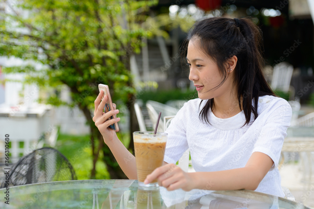 Profile view of young beautiful Asian woman using phone at the coffee shop outdoors Profile view of young beautiful Asian woman using phone at the coffee shop outdoors