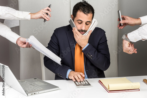 Manager i giacca e cravatta seduto nella postazione di lavoro del suo ufficio è circondato da mani e impegni e fa tante cose contemporaneamente photo