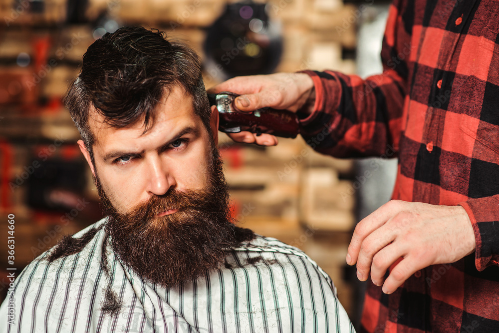 Fototapeta premium Bearded brutal hipster getting hairstyle. Male hairstylist serving client. Barber making haircut using machine and comb. New perfect style.
