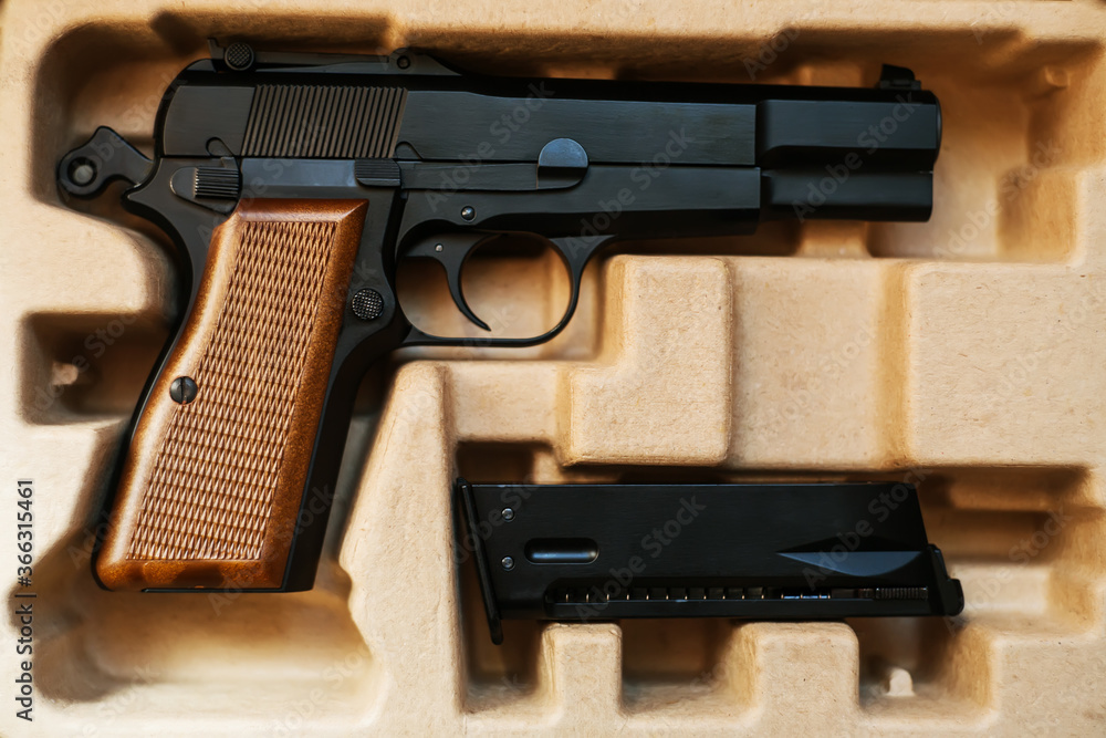 In a cardboard box is an airsoft gun with a brown handle, and next to it is a magazine. Sale and delivery of weapons. In a cardboard box is an airsoft gun with a brown handle, and next to it is a magazine. Sale and delivery of weapons.