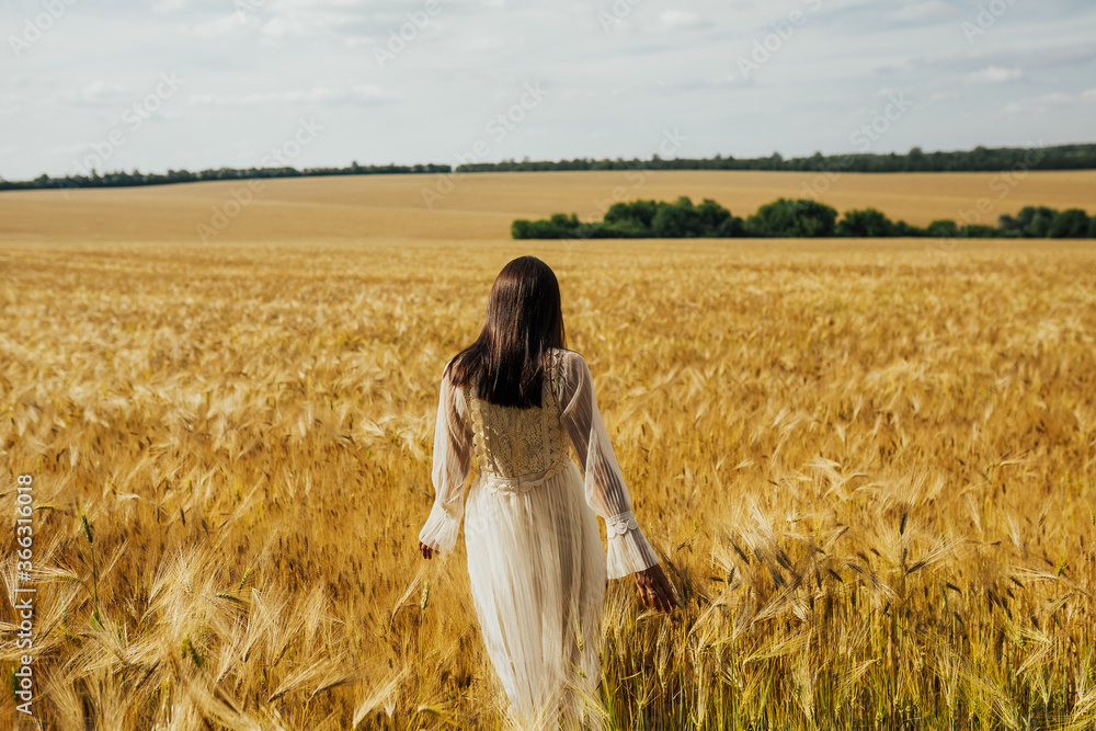 Beautiful young woman in tender white dress walk in wheat field. Photo prom back side. Copy space.