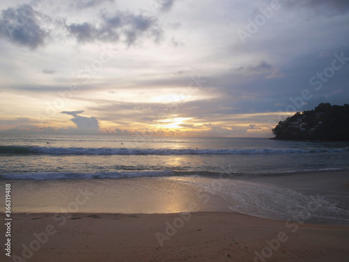 Sunset on the beach and people swimming in the sea. Tropical paradise. Cloudy sky with orange sun. Sundown at the popular resort. Waves, sand, green hill and surfing people. Beautiful heaven with glow