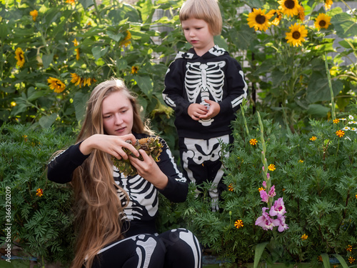 Happy blond boy plays with his sister against backdrop of flower garden with flowers decorated in form of boat. Brother sisiter are dressed in carnival costumes of skeletons. Halloween eve. Close up photo