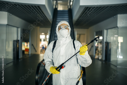 Portrait of a man in a sanitizing disifection suit holding spray near the escalator in an empty shopping mall. A volunteer cleaning up the public places to prevent covid-19. Health awareness concept.