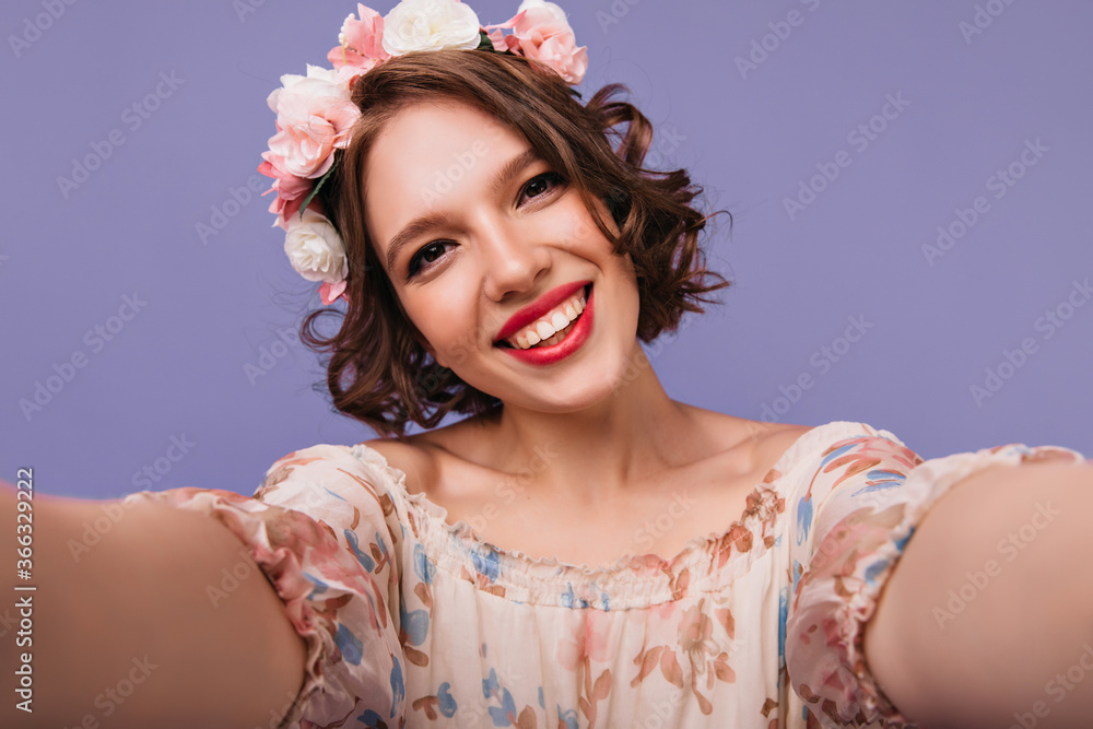 Dark-eyed cute woman in wreath making selfie with smile. Blithesome white girl with short curly hair posing on purple background. Dark-eyed cute woman in wreath making selfie with smile. Blithesome white girl with short curly hair posing on purple background.