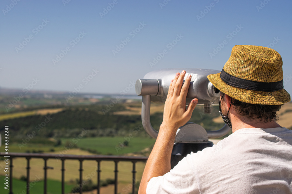 Obraz premium Photo of a young and attractive man wearing a reusable facce mask looking to the nature through some binoculars in a view point. Tourism during coronavirus outbreak