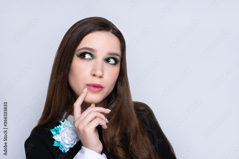Young beautiful girl looks side and touches her lips thoughtfully. Beauty woman skincare concept. Bright makeup green shades shadows sequins. Evening hairstyle long straight hair brunette. Gray back