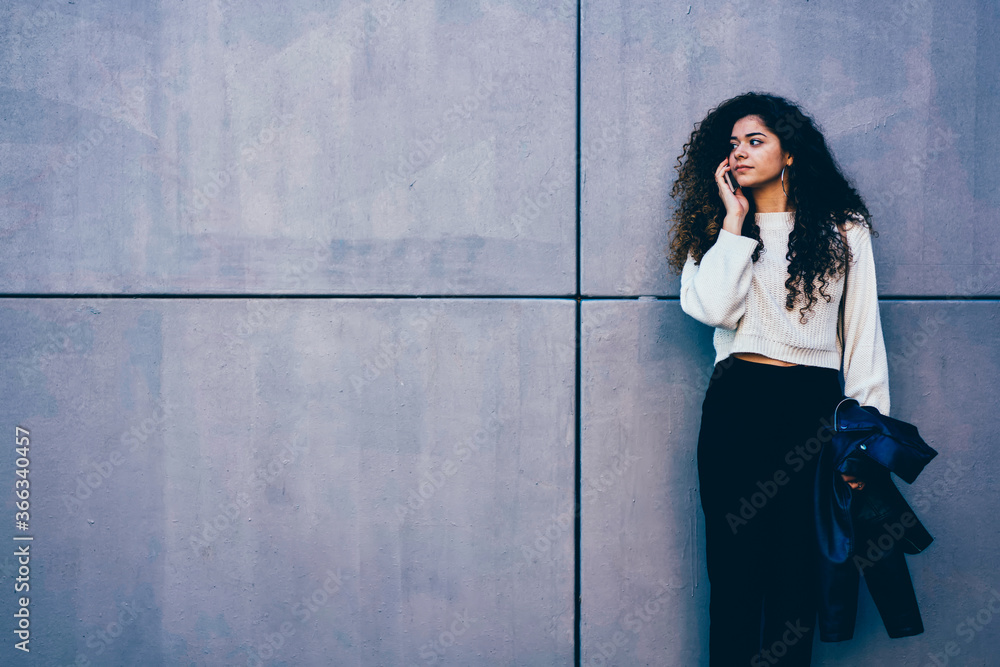 Attractive female student with curly hair having mobile conversation via telephone during spring walk at urban setting.Young pretty woman talking on cellular while strolling near to copy space wall Attractive female student with curly hair having mobile conversation via telephone during spring walk at urban setting.Young pretty woman talking on cellular while strolling near to copy space wall