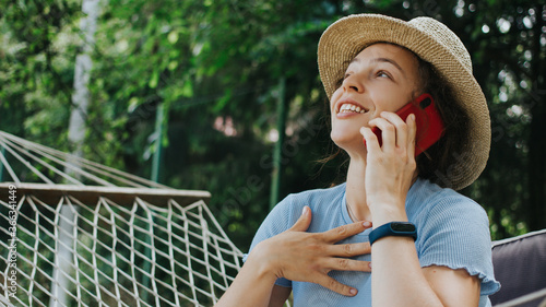 Happy young woman excited about receiving good news by phone conversation.Smiling emotional girl in summer hat talking on smartphone,gesticulate,win,great surprise sitting in hammock in green garden