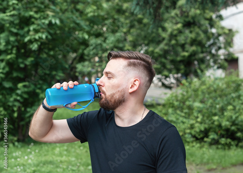 Guy drinks water from a blue bottle. Handsome stylish man after jogging in the park. Drinking water concept