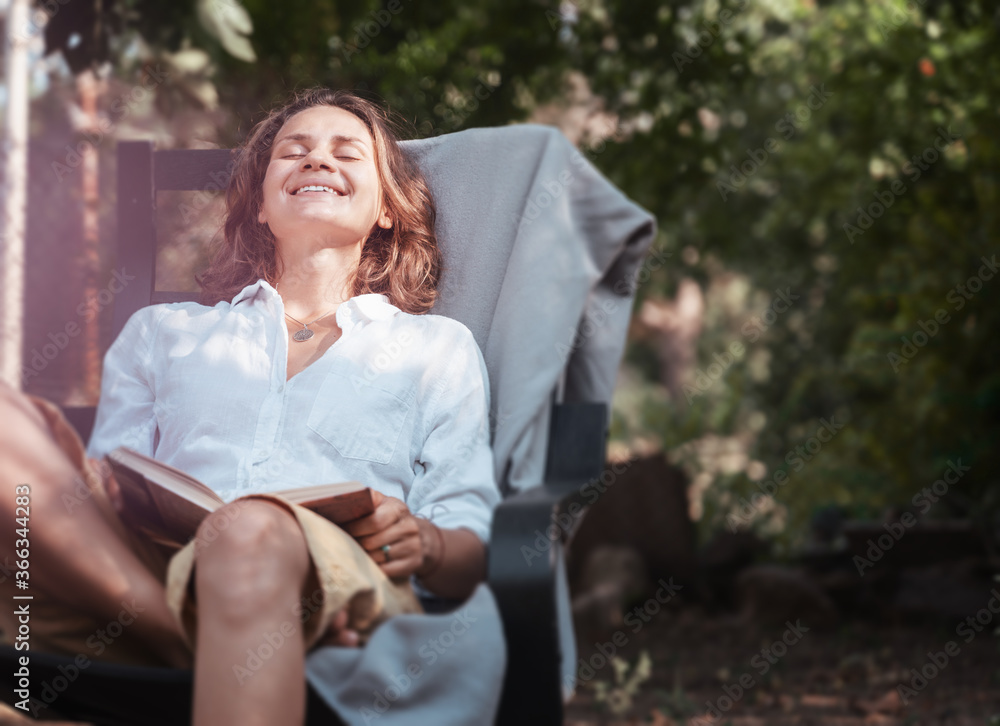 Naklejka premium Young beautiful girl woman enjoys nature and reading a book in the summer in the garden of her house sitting on a chair