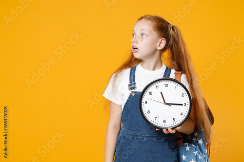 Young redhead school teen kid girl 12-13 years old in white t-shirt blue denim uniform backpack hold in hands clock isolated on yellow background children studio portrait Education lifestyle concept
