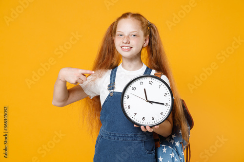 Young redhead school teen kid girl 12-13 years old in white t-shirt blue denim uniform backpack hold in hands clock isolated on yellow background children studio portrait Education lifestyle concept