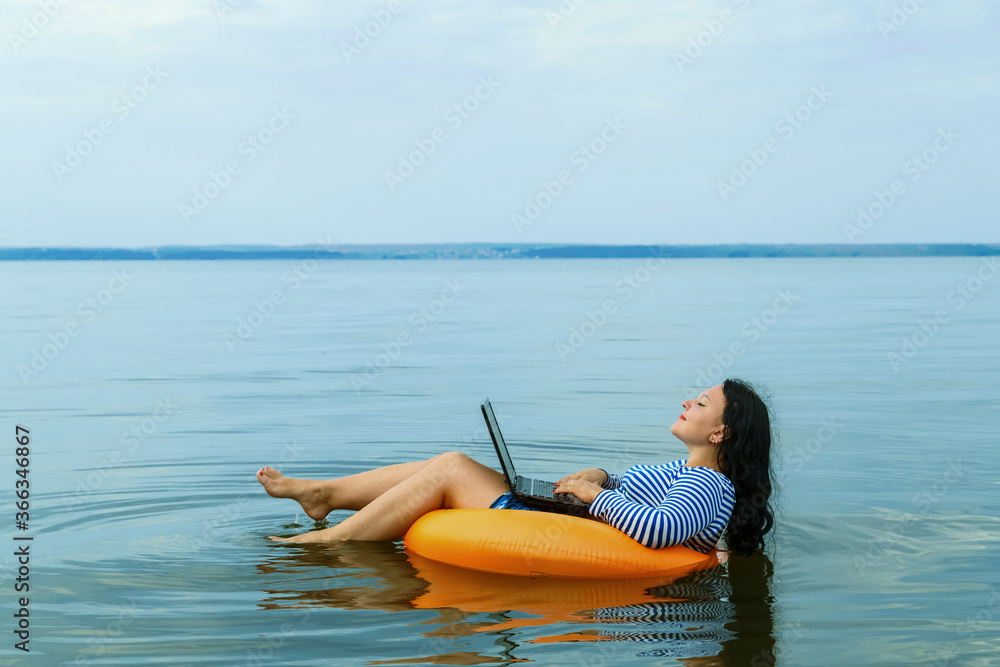 Woman on vacation works remotely with a laptop in a swimming circle in the morning water.