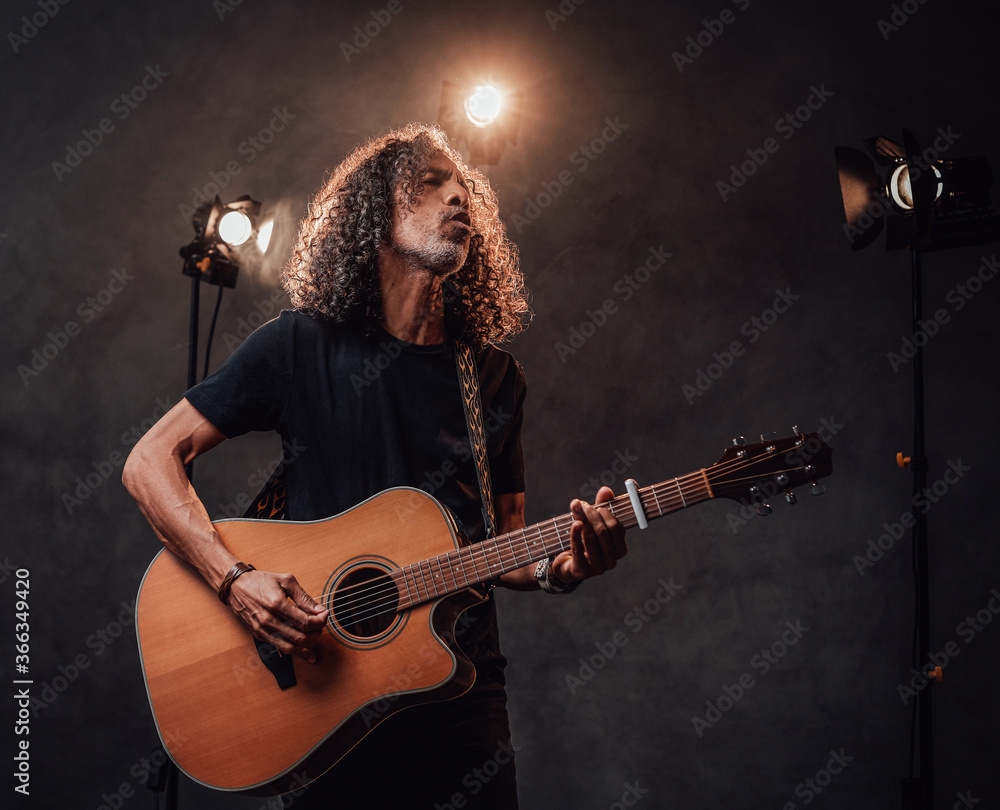 Fototapeta premium Middle aged hispanic musician in black t-shirt emotionally singing and playing guitar. View of musician in the spotlight
