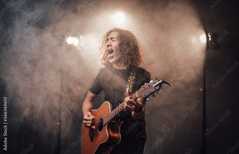 Hispanic musician in black t-shirt emotionally singing and playing guitar in stage lights, surrounded by smoke