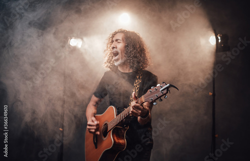 Hispanic musician in black t-shirt emotionally singing and playing guitar in stage lights, surrounded by smoke