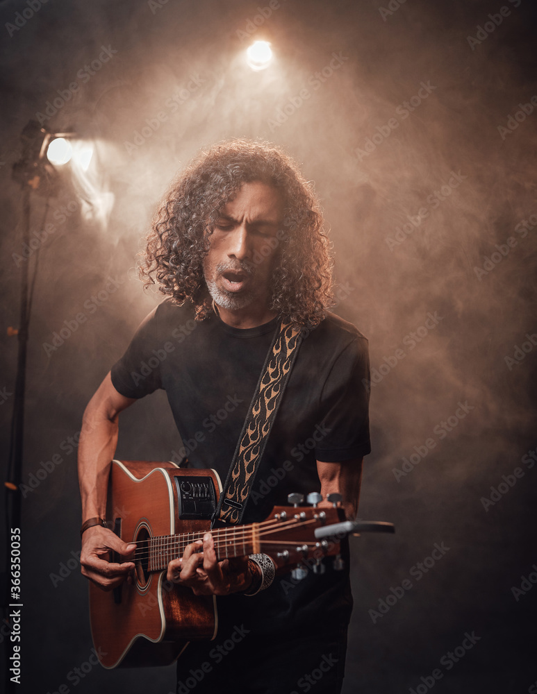 Hispanic musician in black t-shirt emotionally singing and playing guitar in stage lights, surrounded by smoke Hispanic musician in black t-shirt emotionally singing and playing guitar in stage lights, surrounded by smoke