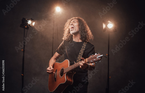 Middle aged hispanic musician in black t-shirt emotionally singing and playing guitar. View of musician in the spotlight