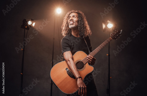Middle aged hispanic man musician in black t-shirt holds guitar. View of musician in the spotlight