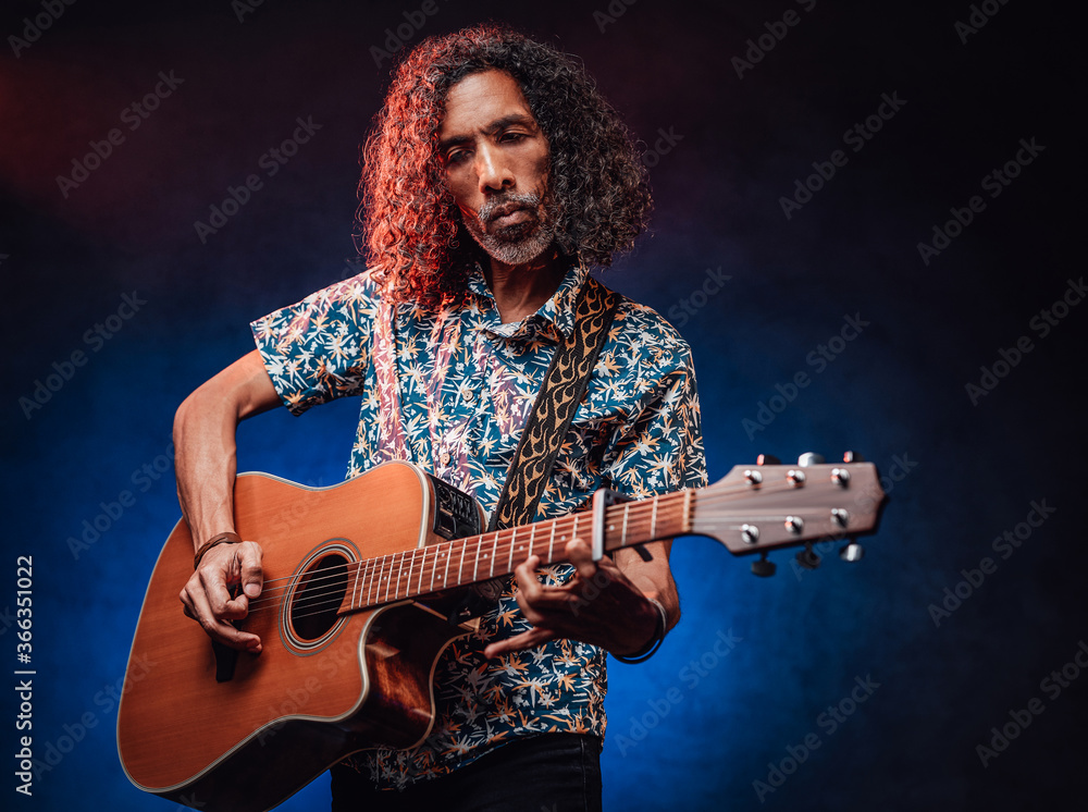 Obraz premium Middle aged hispanic man musician in a hawaiian shirt playing guitar on a dark illuminated by blue and red light. Concept of music, hobby, festival