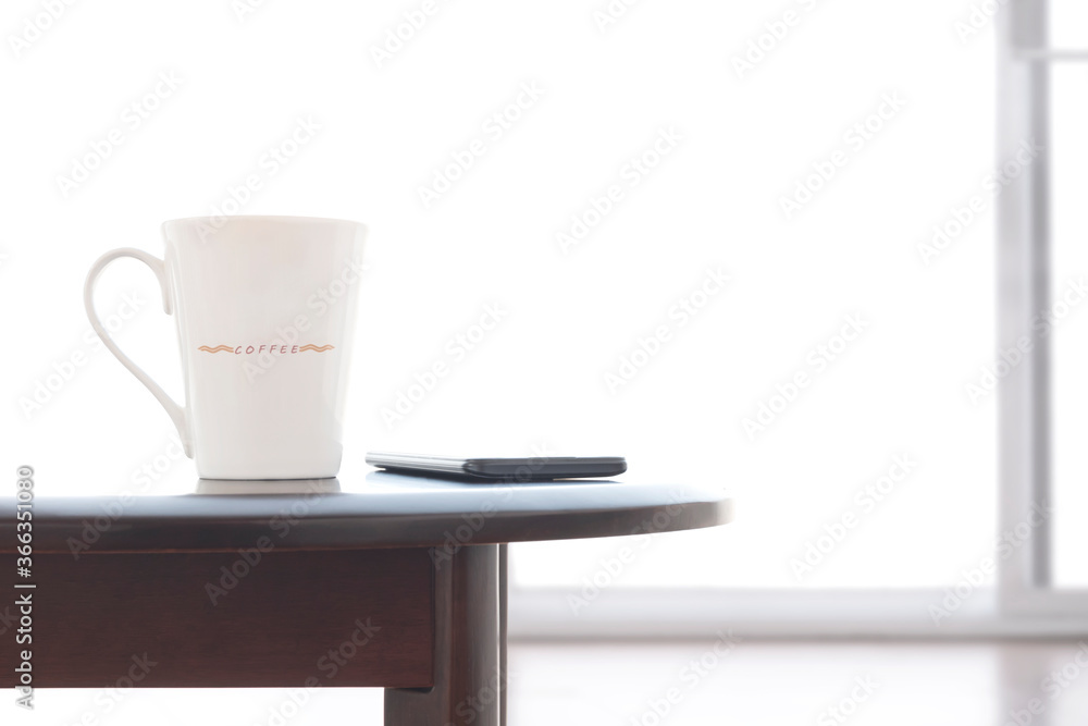 Close up white ceramic coffee cup and smart phone on oval wooden table with blurred frosted glass sliding door in white living room Close up white ceramic coffee cup and smart phone on oval wooden table with blurred frosted glass sliding door in white living room