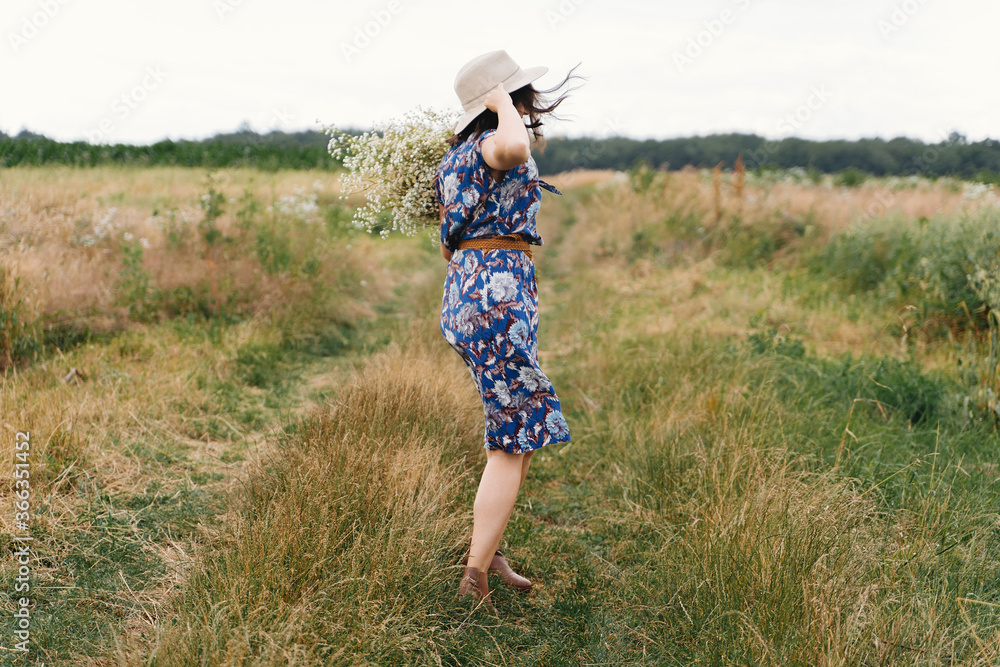 Young woman in blue vintage dress and hat dancing with white wildflowers in meadow. Beautiful girl having fun and laughing in windy field with big daisies bouquet. Authentic summer in countryside