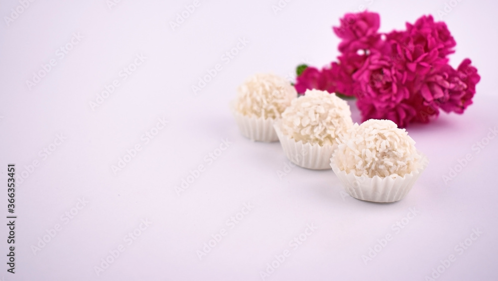 Three white Raffaello balls in coconut flakes with pink little roses on white background. Copy space. Valentine's background. Invitation postcard. 