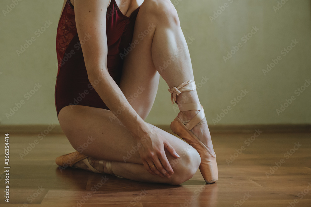 Fototapeta premium Beautiful graceful young ballerina in pointe shoes sitting on the brown wooden floor. No face dancing female feet legs stretching indoors toes professional arts. Ballet practice and stretching