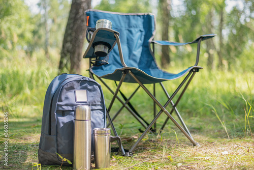 Backpack, vacuum flask, touristic chair and steel cup on the ground on the green forest background.