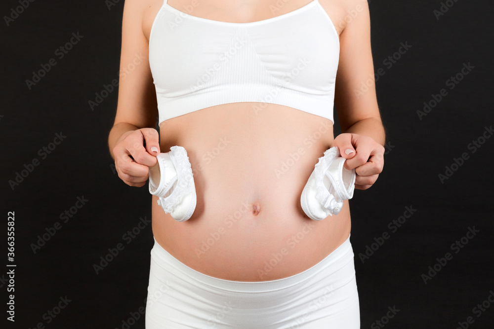 Close up of pregnant woman holding a pair of shoes for a baby girl at black background. Child expecting. Copy space Close up of pregnant woman holding a pair of shoes for a baby girl at black background. Child expecting. Copy space