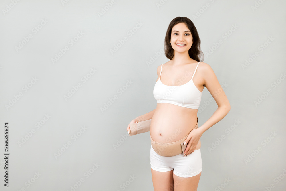 Portrait of pregnant woman in underwear dressing elastic maternity band at gray background with copy space. Orthopedic abdominal support belt concept Portrait of pregnant woman in underwear dressing elastic maternity band at gray background with copy space. Orthopedic abdominal support belt concept