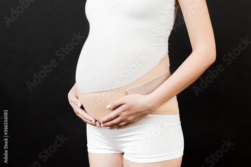 Close up of pregnant woman dressed in orthopedic corset for reducing pain in the back at black background with copy space. Orthopedic abdominal support belt concept © sosiukin