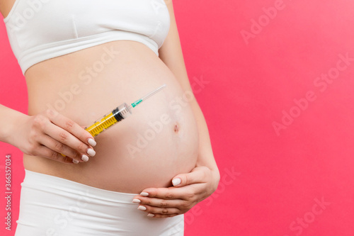Close up of pregnant woman holding a syringe against her belly at colorful background with copy space. Injection during pregnancy concept