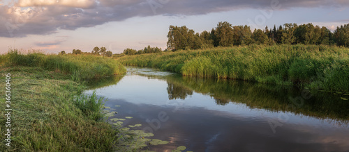 A small river on a green meadow next to a forest reflects sunset clouds