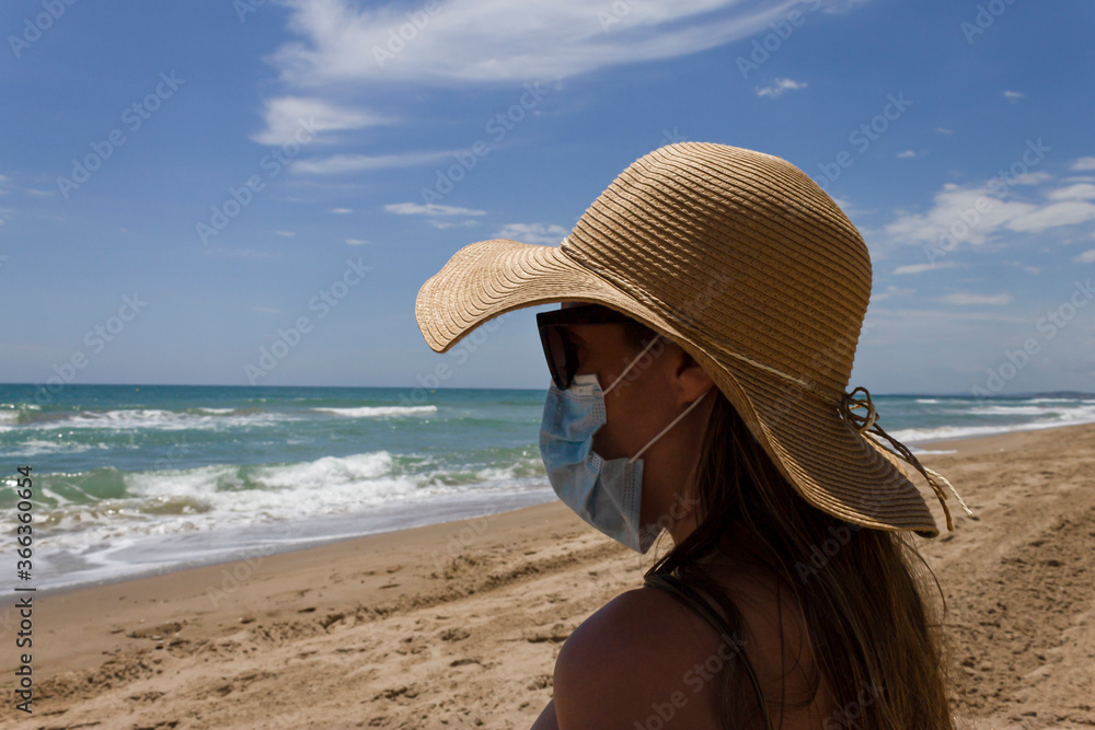 Beautiful young girl sitting on the sand beach wearing straw hat ,sunglasses and medical protection mask ,looking at the coast scenary.Holidays.Coronavirus,quarantine.summer 2020 in Mediterranean sea.