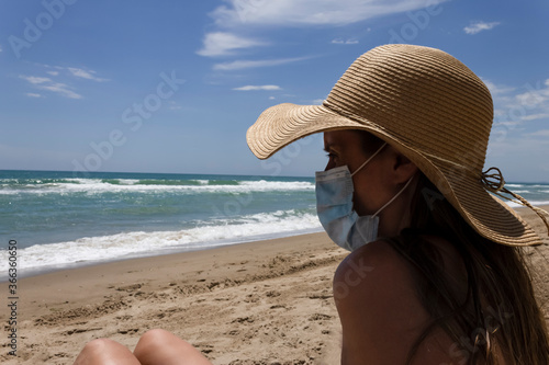 Beautiful young girl sitting on the sand beach wearing straw hat ,sunglasses and medical protection mask ,looking at the coast scenary.Holidays.Coronavirus,quarantine.summer 2020 in Mediterranean sea.