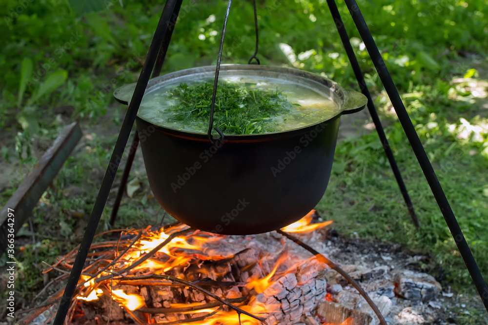 Black cauldron with soup with dill on a tripod over a bonfire Black cauldron with soup with dill on a tripod over a bonfire