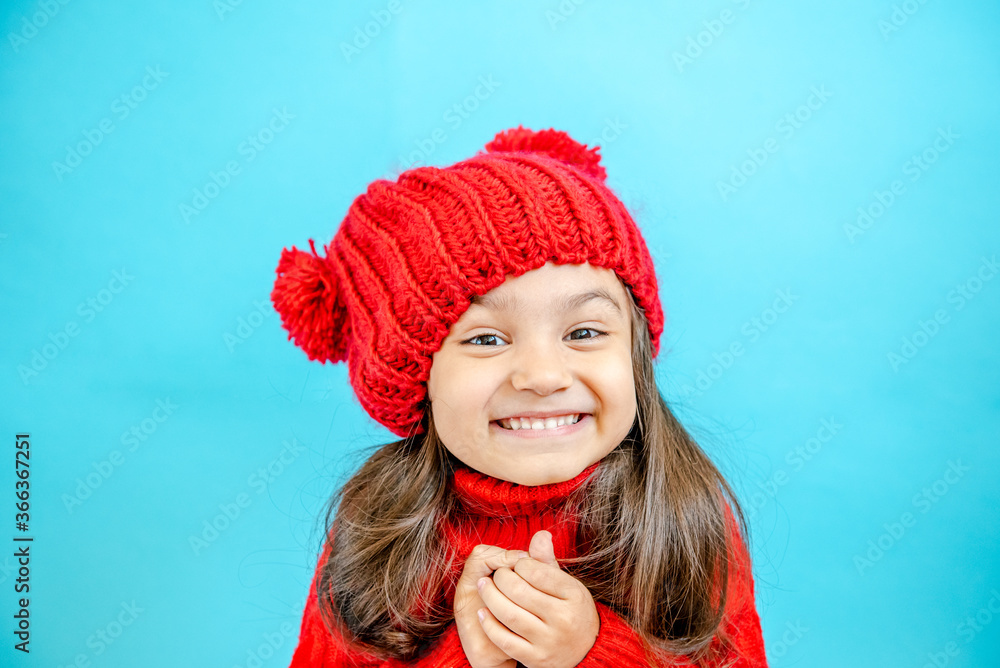 Portrait of a little curly haired girl in a knitted red hat in winter. little girl with dark hair in a red knitted hat and sweater smiling on a blue background isolate, place for text. Winter clothes Portrait of a little curly haired girl in a knitted red hat in winter. little girl with dark hair in a red knitted hat and sweater smiling on a blue background isolate, place for text. Winter clothes