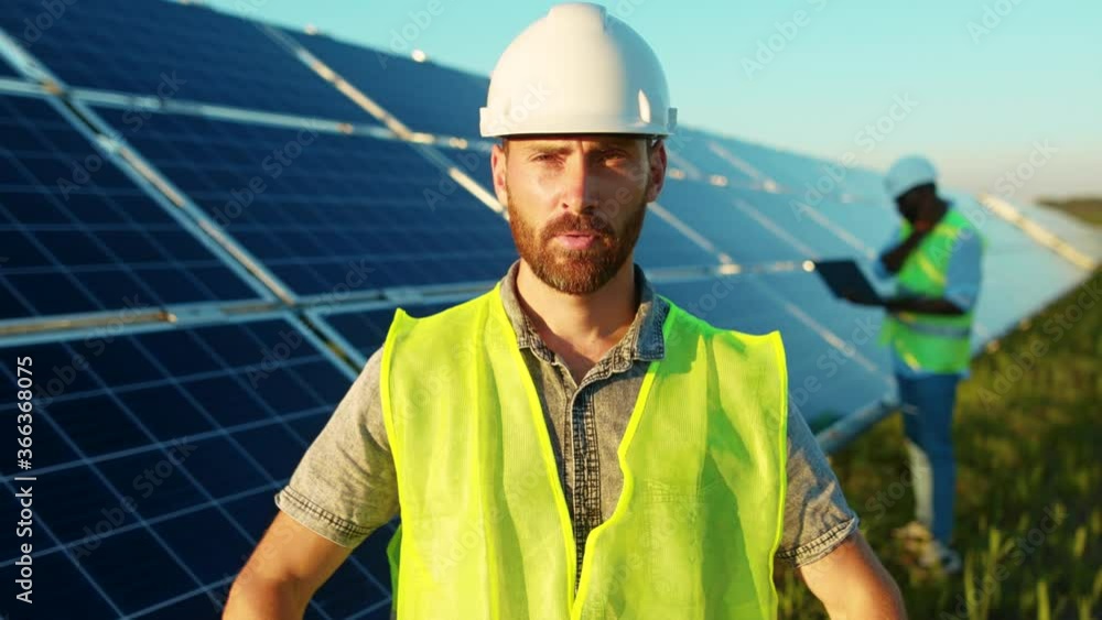 Attractive young worker man stand look at camera near solar batteries construction renewable power at sunlight technology modern electricity energy portrait close up slow motion Attractive young worker man stand look at camera near solar batteries construction renewable power at sunlight technology modern electricity energy portrait close up slow motion
