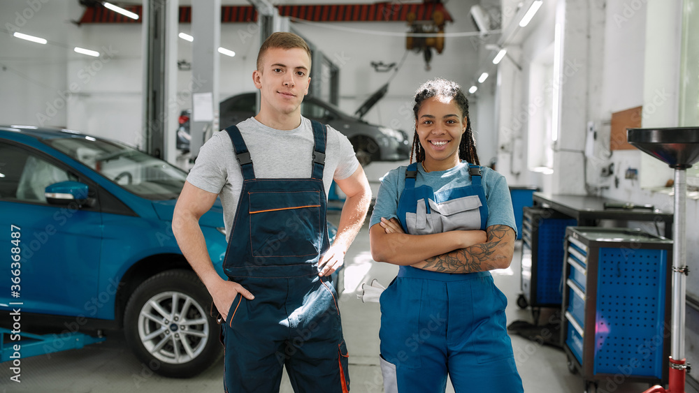 Obraz premium We value your need. Young african american woman and caucasian man, professional female and male mechanic smiling at camera, standing in auto repair shop
