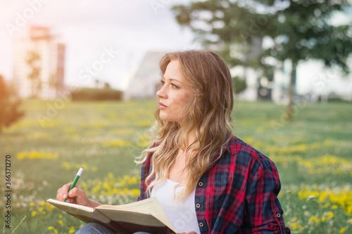 Young caucasian student girl writing notes to notebook while sitting on campus lawn
