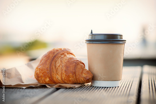 eco paper cup with coffee or tea and croissant on kraft paper, on a wooden table. Breakfast on the street, in the park, on a bench.