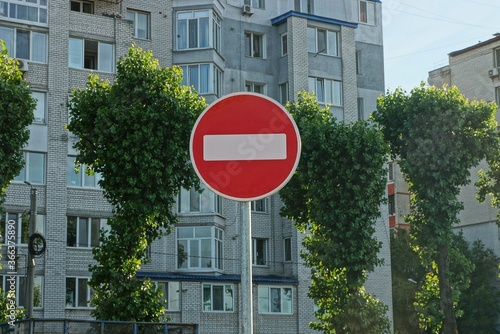 one round road sign no entry on a gray pillar against the background of green trees and a multi-storey building