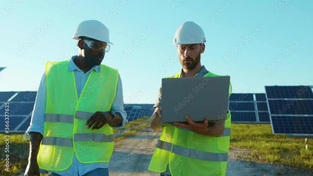 Slow motion african american and caucasian engineers walking with laptop on the solar farm in special uniform discuss the installation of sunny batteries electricity environmental Slow motion african american and caucasian engineers walking with laptop on the solar farm in special uniform discuss the installation of sunny batteries electricity environmental
