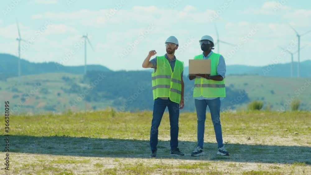 Close up two young engineers use laptop stand near windmills in fields team work discuss labor collaboration contribution windpower farm technicians talking slow motion Close up two young engineers use laptop stand near windmills in fields team work discuss labor collaboration contribution windpower farm technicians talking slow motion