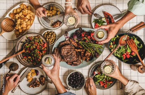 Summer barbeque party. Flat-lay of table with grilled meat, vegetables, salad, roasted potato and peoples hands with glasses of lemon water over white tablecloth, top view. Family gathering concept